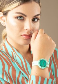 Un reloj turquesa con una esfera redonda y correa de silicona se lleva en la muñeca de una modelo. La modelo lleva una blusa a rayas en tonos verde azulado y marrón.
