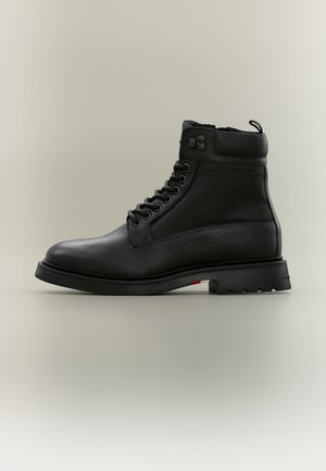 Bottine noire en cuir et tissu à lacets avec semelle épaisse et languette sur fond clair uni.