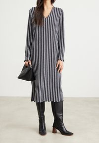 Robe longue rayée en bleu marine et blanc, avec un col en V, des manches longues et une silhouette fluide. Portée avec des bottes noires montantes et un sac noir.