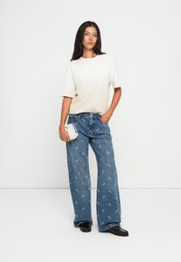 Kermainen lyhythihainen paita, jossa on brodeerattu teksti, yhdistettynä vaaleansinisiin leveisiin farkkuihin, joissa on toistuva valkoinen logokuvio. Mustat kengät ja valkoinen laukku.