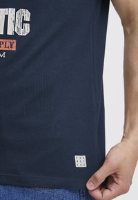 Námornícky modré bavlnené tričko s bielym a oranžovým potlačeným textom; na spodnom leme je malá nášivka s nápisom "DNM WRKRS". Hladká textúra.