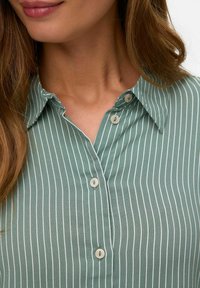 Chemise verte à manches courtes avec des rayures verticales blanches, dotée d'un col et de deux boutons visibles au niveau du cou, fabriquée dans un tissu lisse.