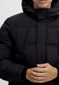 Black puffer jacket with a high collar, featuring a zip closure, padded texture, and side pockets. Ribbed cuffs complete the design.