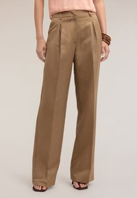 Pantaloni a gamba larga marroni realizzati in un tessuto liscio, con pieghe frontali e una vestibilità sartoriale, abbinati a una maglietta rosa chiaro e sandali con cinturini.