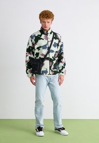 Giacca in pile verde, rosa e blu con motivo mimetico, dotata di colletto alto; abbinata a jeans azzurri chiari e una borsa a tracolla nera.
