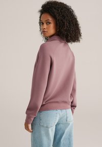 Roza majica dugih rukava s visokim ovratnikom i rebrastim manžetnama, uparena sa svijetloplavim trapericama. Tkanina izgleda mekano i opuštenog kroja.