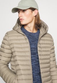 Beige padded jacket with a hood, features horizontal stitching, worn over a textured blue sweater. Light green cap completes the outfit.