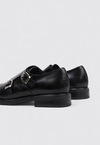 Zapatos de cuero negros con punta redondeada, que cuentan con un acento de hebilla plateada y un tacón bajo tipo apilado. Textura suave y diseño minimalista.