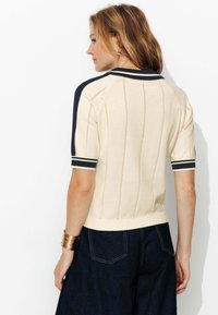 Pull en tricot à manches courtes beige avec des accents navy, col et poignets côtelés, présentant des détails de couture verticaux pour une texture.