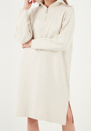 Photo recadrée d’un mannequin portant une robe en maille couleur crème, avec un col zippé et une fente latérale. Elle tient une main près de son visage devant un fond uni.