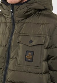 Giacca puffer verde oliva con chiusura a zip, cappuccio regolabile e tasca frontale con patta che presenta un logo a patch nero. Texture liscia e lucida.