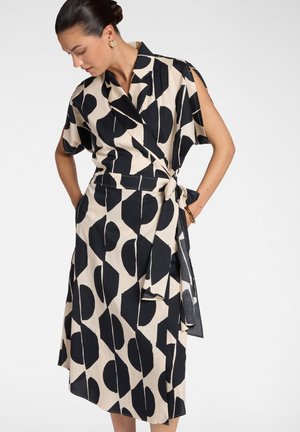 Donna che indossa un vestito avvolgente beige con grandi forme astratte nere, maniche corte e cintura annodata in vita, guarda verso il basso con le mani nelle tasche.