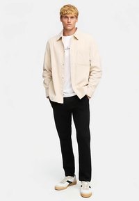 Veste-chemise en velours côtelé beige portée sur un t-shirt blanc, pantalon noir et baskets blanches avec des accents marron clair. Design minimaliste, poche avant visible.