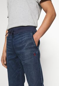 En person som bär mörkblå denim-joggers med dragsko i midjan och röd broderad logotyp, handen i höger ficka, samt en ljusgrå t-shirt.