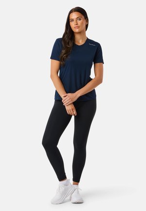 Frau mit langen dunklen Haaren, die ein dunkelblaues kurzärmeliges Shirt, schwarze Leggings und weiße Turnschuhe trägt, steht mit verschränkten Armen.