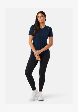 Frau mit langen dunklen Haaren, die ein dunkelblaues kurzärmeliges Shirt, schwarze Leggings und weiße Turnschuhe trägt, steht mit verschränkten Armen.