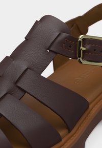 Καφέ δερμάτινο σανδάλι με φαρδιές ράβδους, μια χρυσή αγκράφα και ταμπά επενδεδυμένη σόλα σε λευκό φόντο.