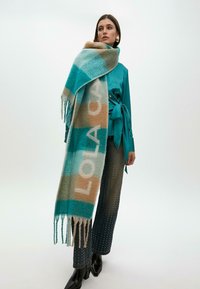 Tyrkysový a béžový kostkovaný šátek s třásněmi, na kterém je velkými písmeny nápis "LOLA". Modelka má na sobě tyrkysový saténový top a tmavé zdobené kalhoty.