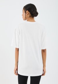 Volný, krátký bílý tričko s krátkým rukávem z měkké bavlny. Má zaoblený výstřih a mírně delší zadní lem. Minimalistický design.
