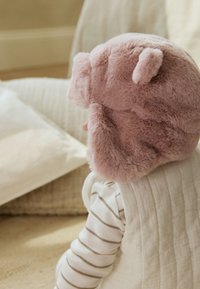 Rožinės spalvos minkštas kailio gyvūno kepurė suapvalintomis ausimis, minkštu tekstūra ir prigludusiu prigludimu. Dėvima kartu su dryžuotu ilgomis rankovėmis marškinėliu ir siuvinėta liemene.