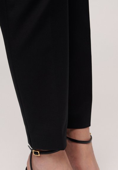 Zwarte tapered broeken in een gladde stof met een slim fit silhouet, featuring een gouden vierkante accent aan de zoom en dunne zwarte enkelbandjes.