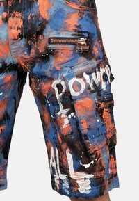 Shorts cargo pour hommes avec un motif de taches de peinture bleues, orange et noires, dotés de zippers et d'un texte de style graffiti blanc sur la poche de la jambe.