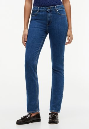 Femeie purtând blugi bleumarin slim și pantofi loafer din piele neagră cu ciucuri, stând în picioare pe un fundal alb.