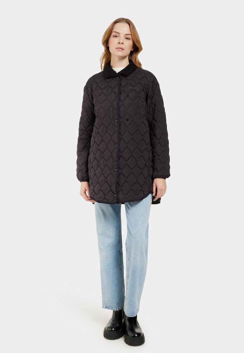 Schwarze gesteppte Jacke mit einem strukturierten Rautenmuster, Knopfverschluss und einem weichen Kragen. Getragen mit hellblauen Jeans und schwarzen Stiefeln.