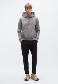 Hall halliga kapuutsiga dressipluus reljeefse "BOSS" logoga, kängurutasku, soonikkoes varrukaotsad, mustade kitsenevate pükstega ja beežide tossudega.