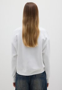 Persoon met lang ombre-haar, gekleed in een witte sweatshirt en blauwe jeans, staande tegen een effen lichtgrijze muur.