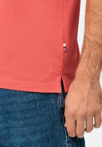 Chemise polo rouge avec une finition texturée, présentant des fentes sur les côtés et une petite étiquette multicolore en bas. Associée à un denim bleu foncé.