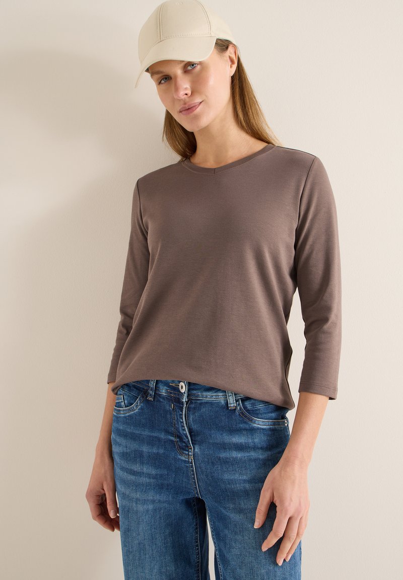 Chemise marron à manches longues avec col en V, associée à un jean en denim bleu. Le mannequin porte une casquette de couleur claire. Matière douce, coupe décontractée.