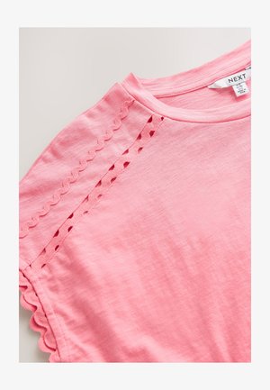 Ružové tričko s krátkym rukávom a okrúhlym výstrihom, zdobené vlnkovanými okrajmi a výrezmi pozdĺž švov na ramenách. Materiál z bavlny.