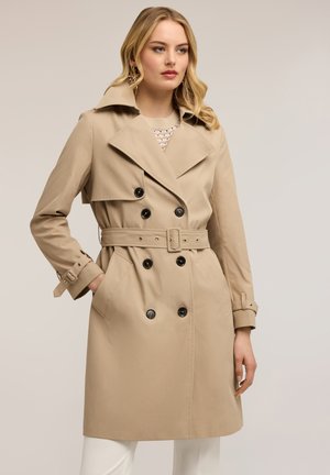 Donna bionda che indossa un trench beige doppiopetto con cintura e bottoni neri, mani nelle tasche, in piedi su uno sfondo neutro.