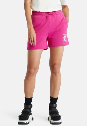 Femme portant un short rose vif à cordon avec le logo Timberland, des chaussettes noires et des sandales noires à semelle blanche, debout les mains près des poches.