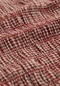 Tessuto a quadretti rossi e crema, con trama arricciata e cuciture arruffate. Il materiale sembra leggero e presenta un motivo a vichy.