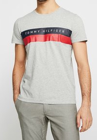 Harmaa puuvillapaita, jossa on pyöreä pääntie. Paidassa on vaakasuora laivastonsininen ja punainen raita rinnassa, jossa lukee "TOMMY HILFIGER".