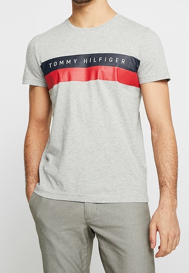 Harmaa puuvillapaita, jossa on pyöreä pääntie. Paidassa on vaakasuora laivastonsininen ja punainen raita rinnassa, jossa lukee "TOMMY HILFIGER".