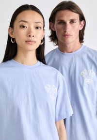 Hellblaue Baumwoll-T-Shirts mit kurzen Ärmeln, verziert mit weißen grafischen Designs auf der Brust, getragen von einer Frau und einem Mann.