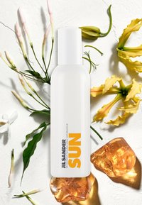 Botella de spray blanca con una etiqueta amarilla que dice "SOL". Rodeada de hojas verdes y flores amarillas sobre un fondo blanco texturizado.