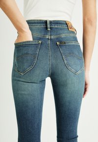 Niebieskie dżinsy o dopasowanym kroju, średnim stanie, tylne kieszenie z zaokrąglonymi szwami oraz skórzana metka przy pasku.