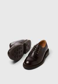 Scarpe in pelle marrone con punta arrotondata, design classico con lacci, suola in gomma con tasselli circolari e cuciture dettagliate lungo le linee.