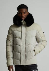 Lys beige puffjakke med svart faux pelskrave. Har en kviltet tekstur, glidelås foran og to sidelommer.
