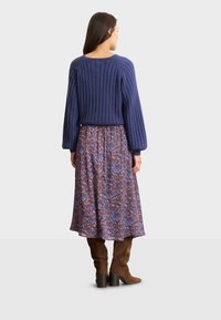 Knitmarineblauer Pullover mit geribbelter Textur, kombiniert mit einem gemusterten, fließenden braunen Kleid mit blauen und roten Akzenten sowie braunen kniehohen Stiefeln.