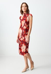 Abito plissettato floreale rosso con scollo alto e maniche a cappuccio. Presenta una miscela di tonalità di rosso profondo e beige chiaro. Indossato con scarpe col tacco nude.