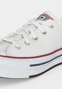 Λευκό χαμηλό καμβά Converse sneaker με λευκά κορδόνια, κόκκινες και μπλε ρίγες στη σόλα και ένα λογότυπο στη γλώσσα.