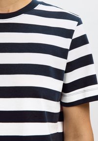 Personne portant une chemise à manches courtes avec des rayures horizontales noires et blanches sur un fond clair uni.