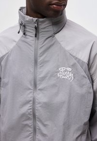 Muž nosící světle šedou bundu na zip s vysokým límcem a bílým vyšitým nápisem na hrudi „Karma“ s očima nad ním.