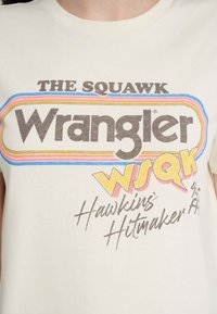Baumwoll-T-Shirt in Cremefarbe mit retro-inspiriertem Text in Braun, Blau und Orange, auf dem "Wrangler" und "WSQK" deutlich zu sehen sind.