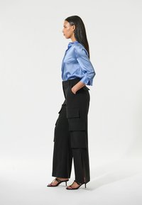Satin blauwe blouse met lange mouwen, gecombineerd met zwarte cargo pants met grote zakken en zijsplitten, gedragen met zwarte enkelbandjes.